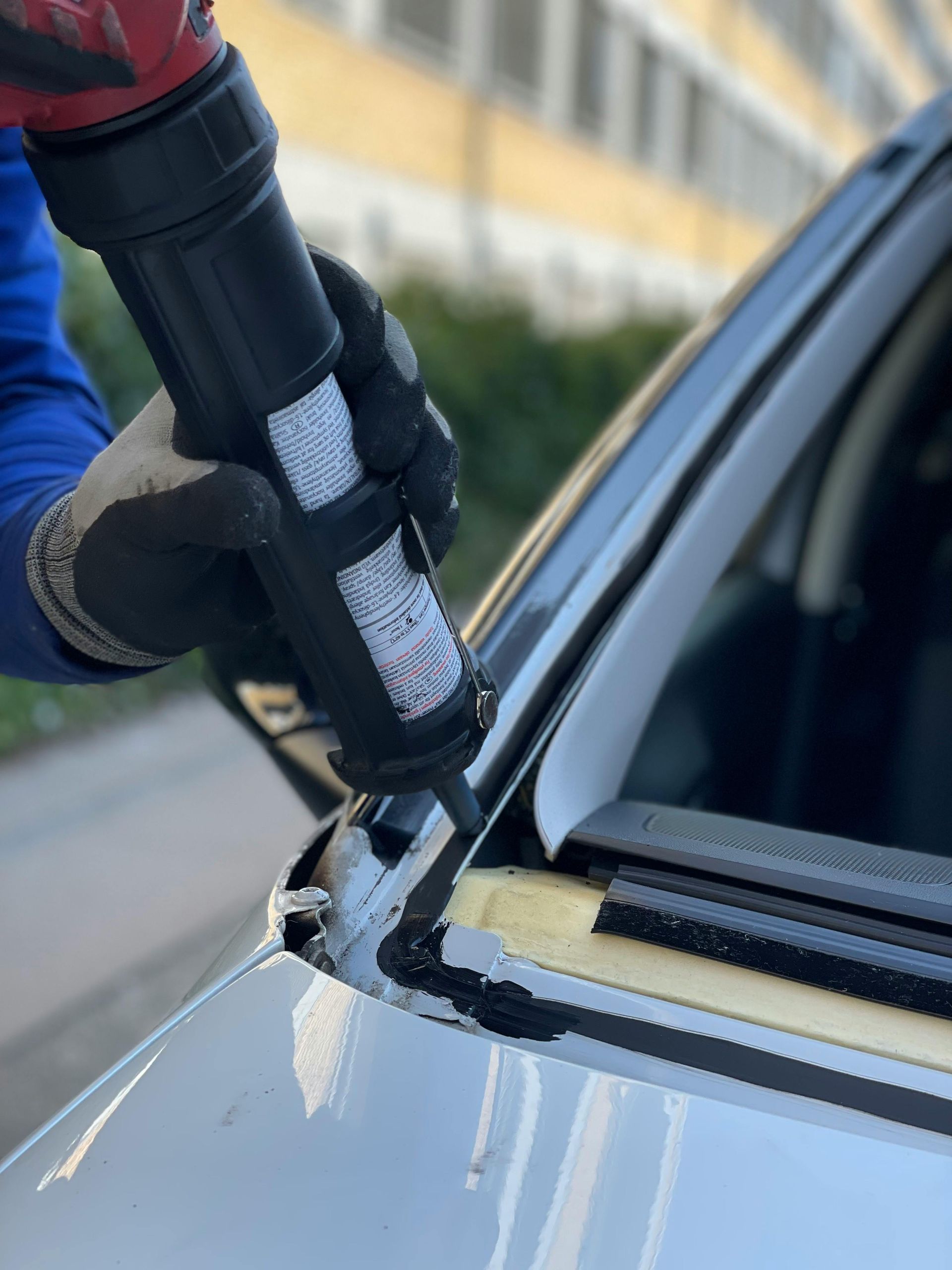 Gloved worker applying black sealant to a car windshield edge with a caulking gun