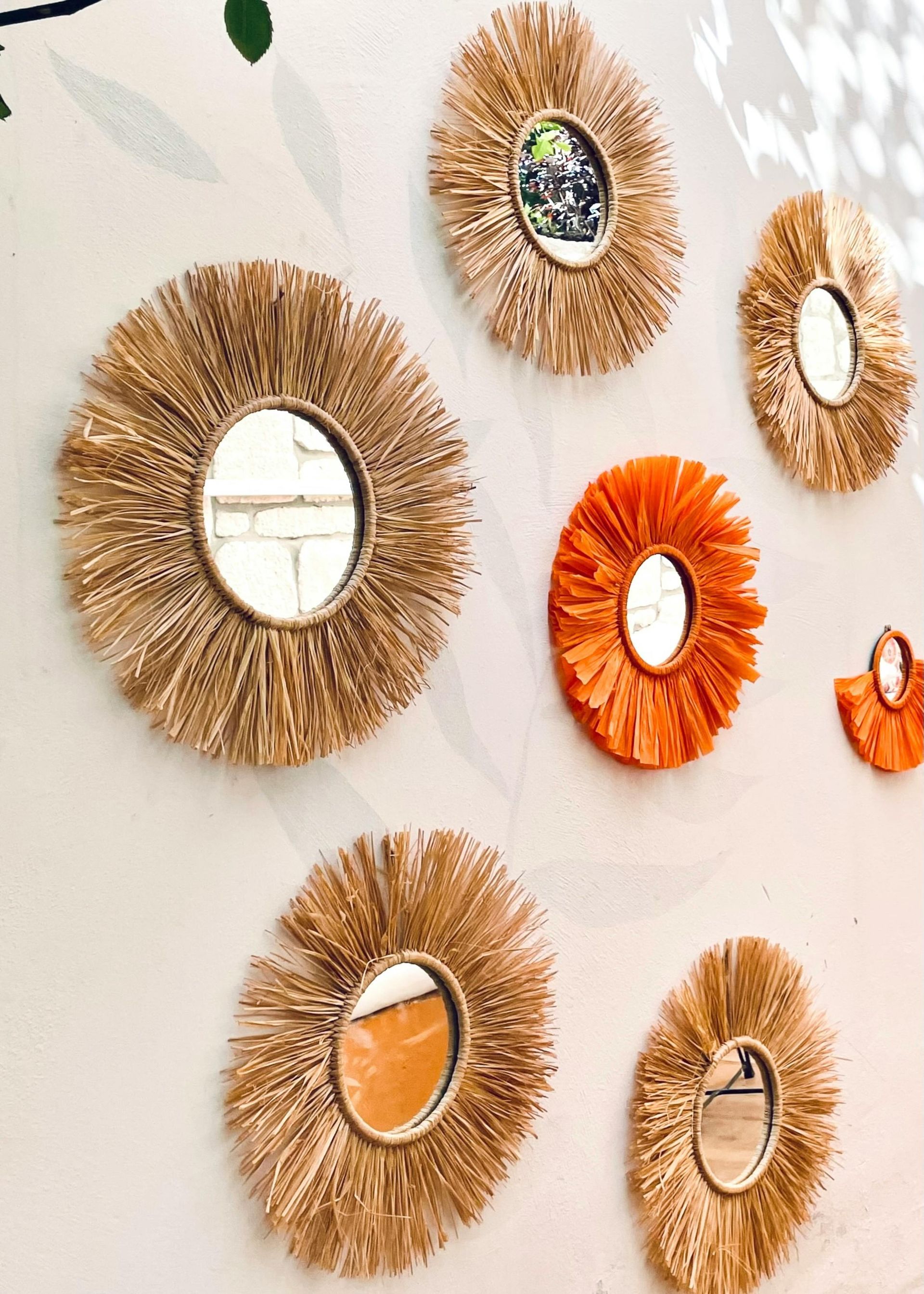 Cluster of decorative straw-framed mirrors and wall art in beige, orange, and gold on a light wall