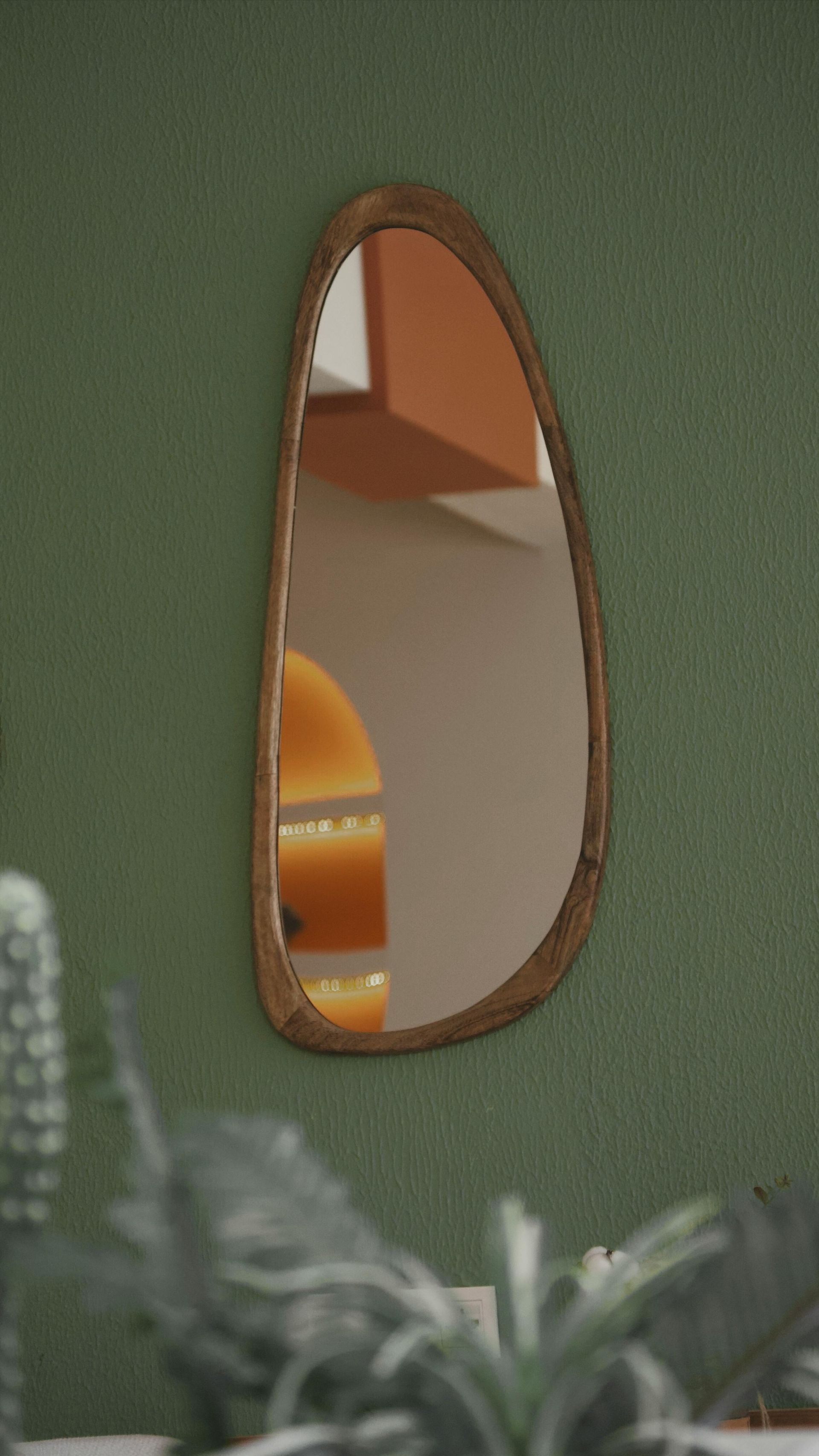 Teardrop-shaped wooden mirror on a green wall, reflecting a warm orange lamp and part of a room.