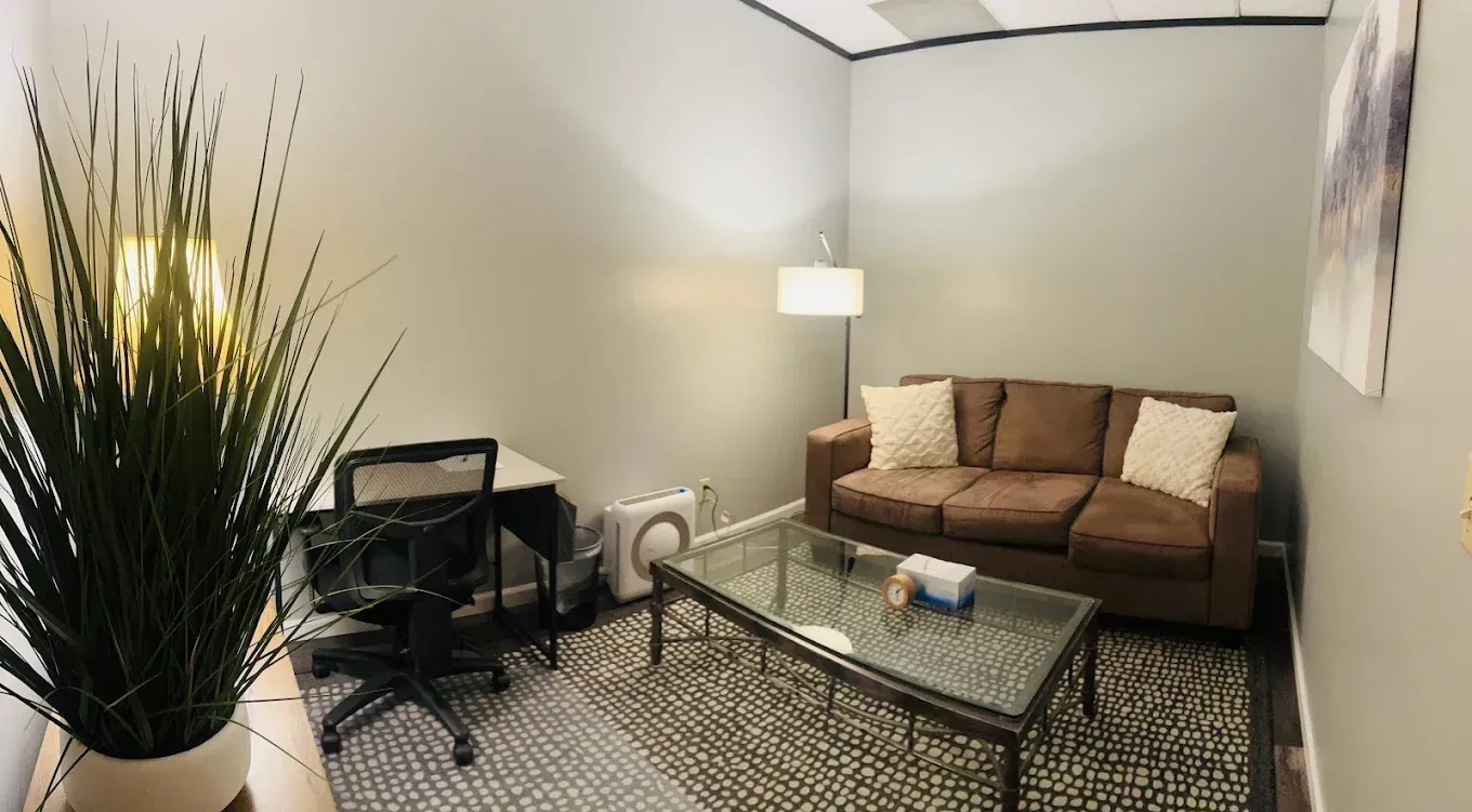 Modern living room with brown sofa, glass coffee table, lamp, chair, and tall plant
