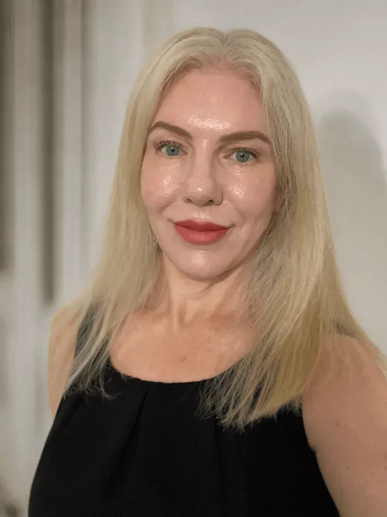Blonde woman in a black sleeveless top, smiling against a neutral indoor backdrop
