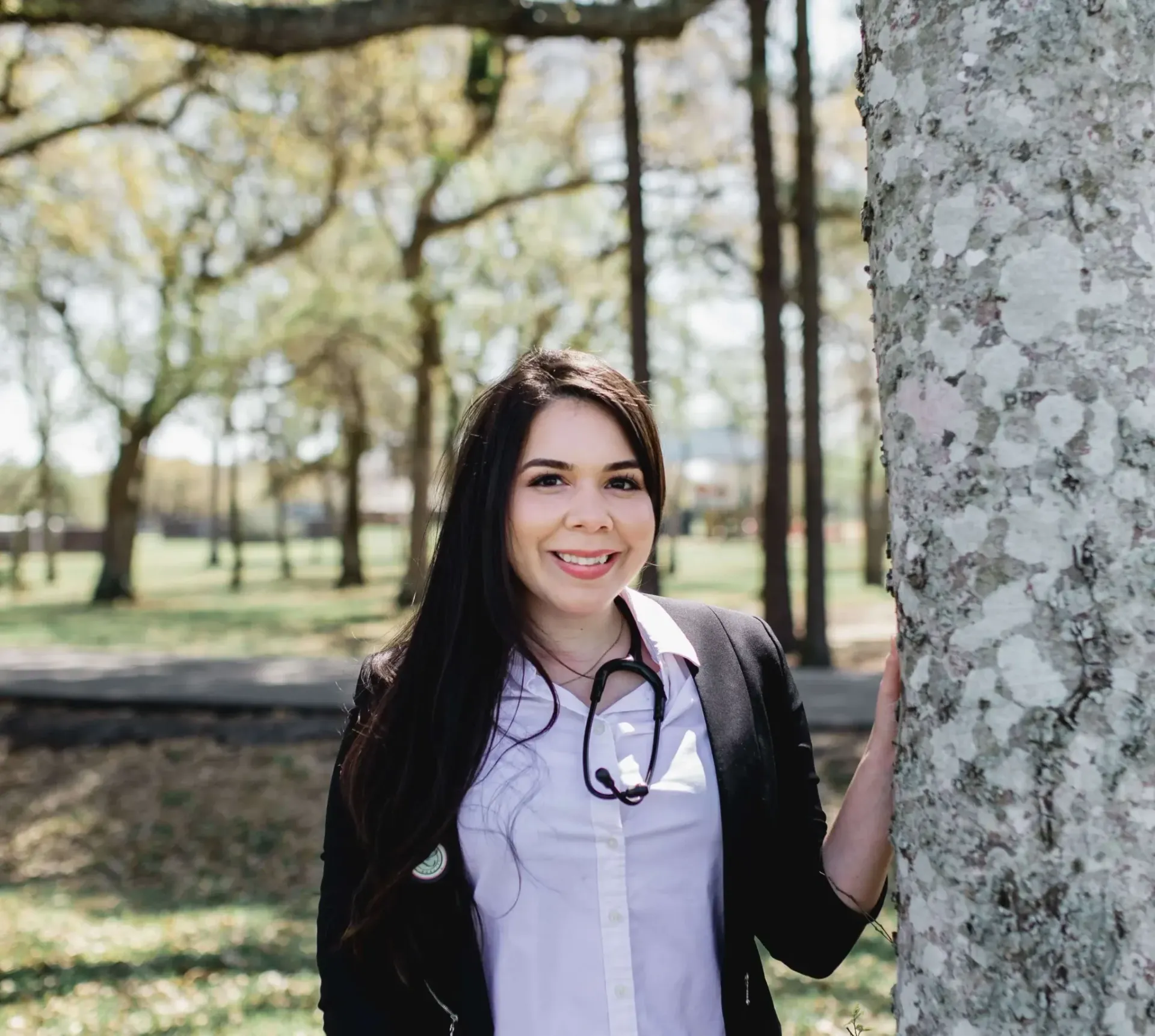 Woman in a black cardigan and white blouse smiling beside a tree in a sunny park