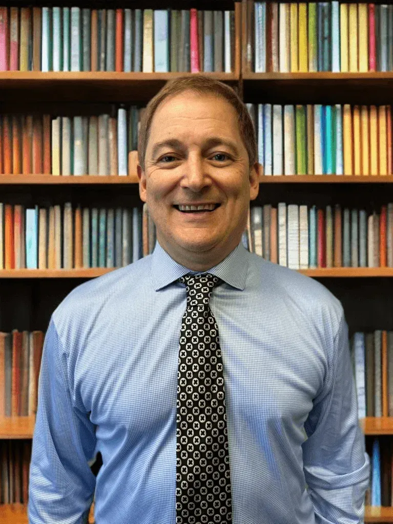 Smiling man in a blue shirt and patterned tie standing before a bookshelf backdrop