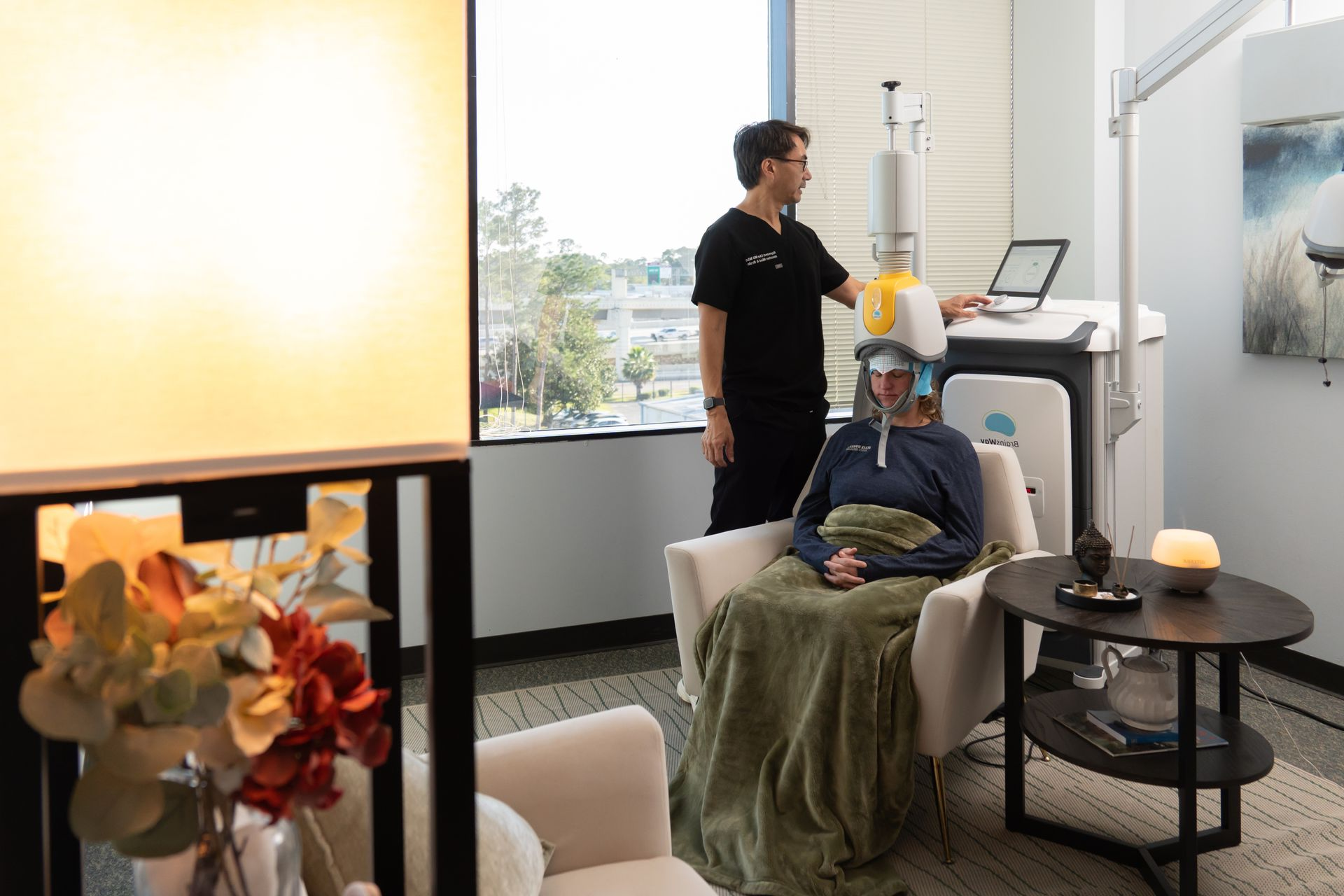 Therapy session in a bright room, with a person in a chair wearing a headset and a clinician standing nearby.