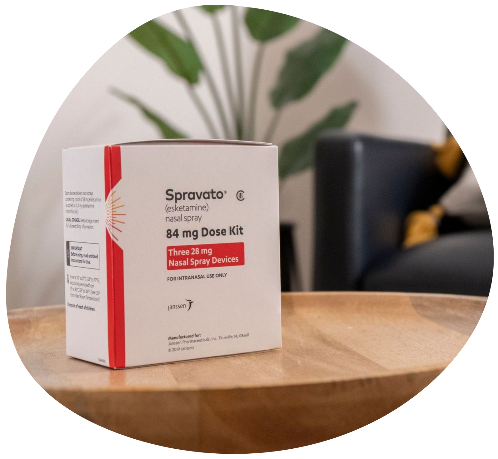 Spravato nasal spray box on a wooden table, with a plant and chair blurred in the background