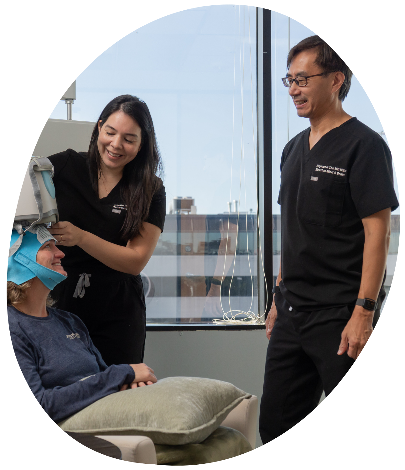 Two clinicians smiling with a seated patient in a bright hospital room near a window.