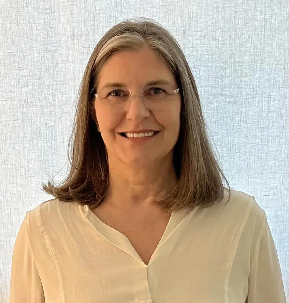 Smiling person with shoulder-length hair and glasses in a cream blouse against a light textured background