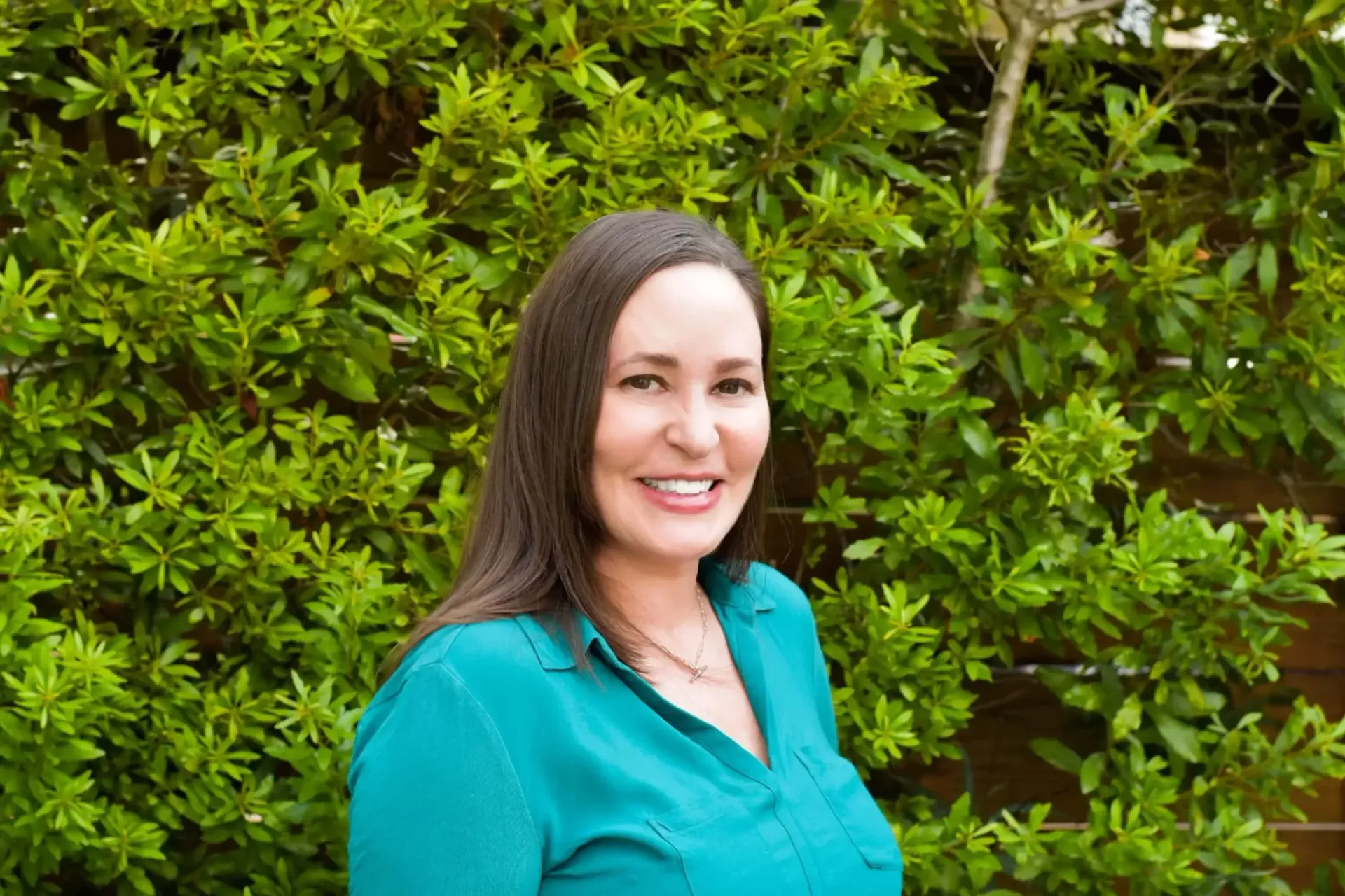 Smiling woman in a teal shirt standing in front of green foliage