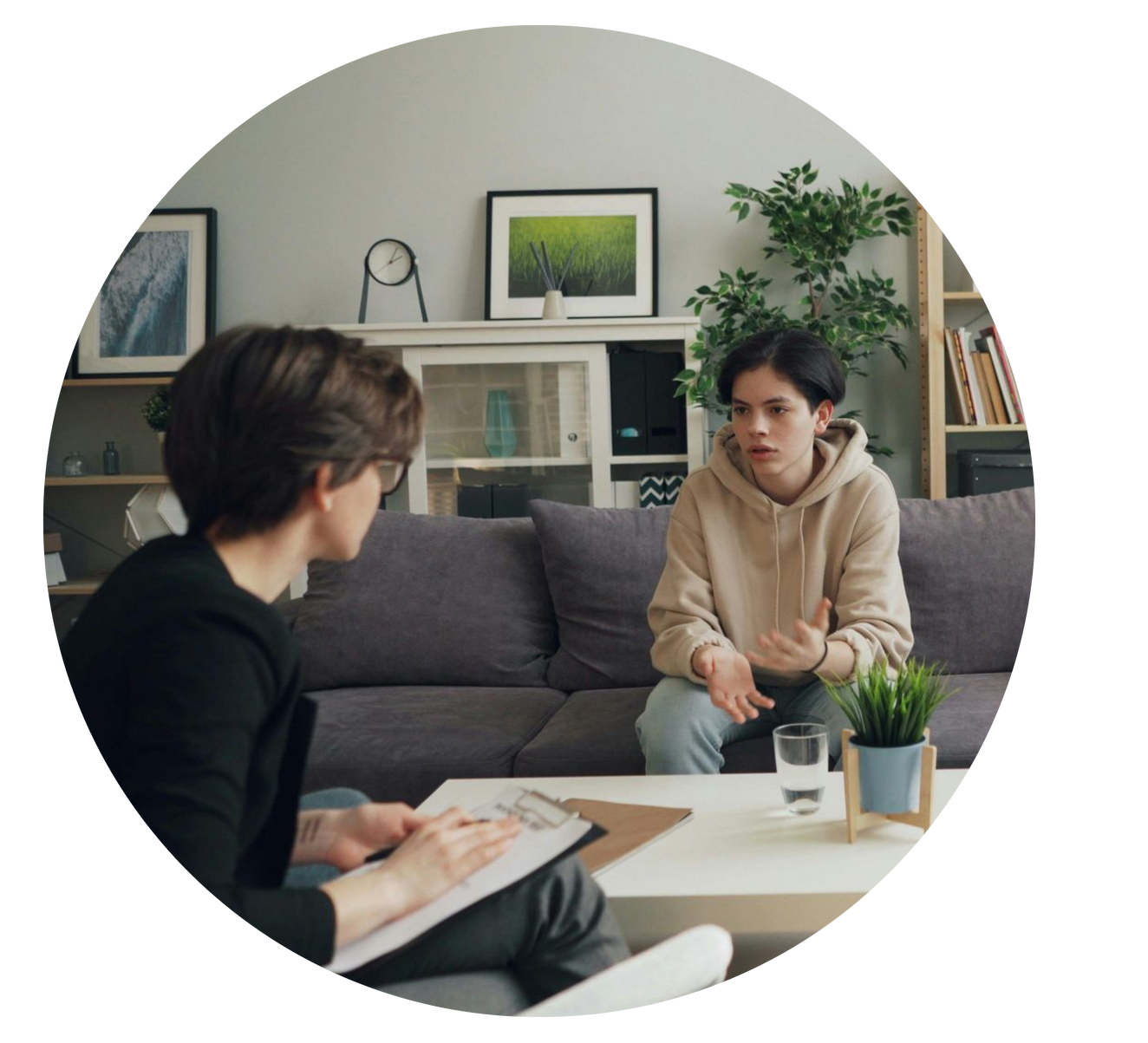 Two people talking across a coffee table in a cozy living room, with a sofa and framed art behind them.