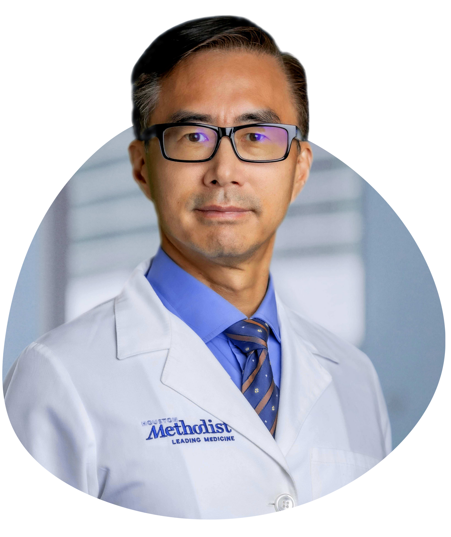 Portrait of a doctor in a white coat and blue tie, wearing glasses, against a blurred clinical background