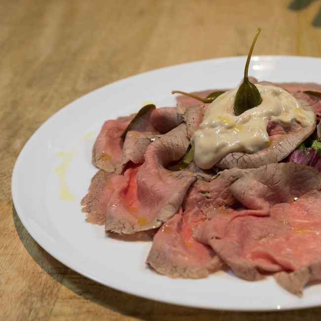 Roast beef tagliato sottile su un piatto bianco, condito con salsa e capperi; su un tavolo di legno.