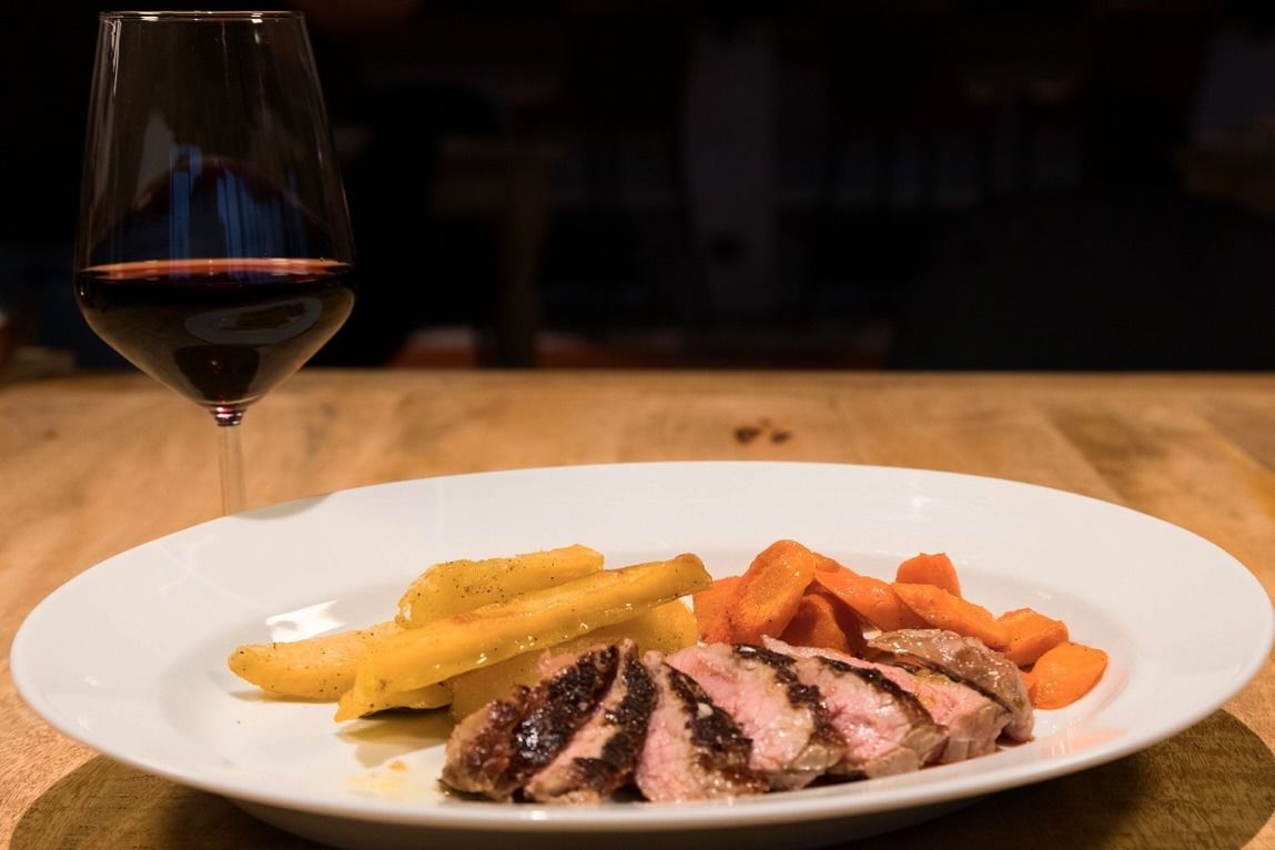 Piatto di carne affettata con carote e patate arrostite, accompagnato da un bicchiere di vino rosso.