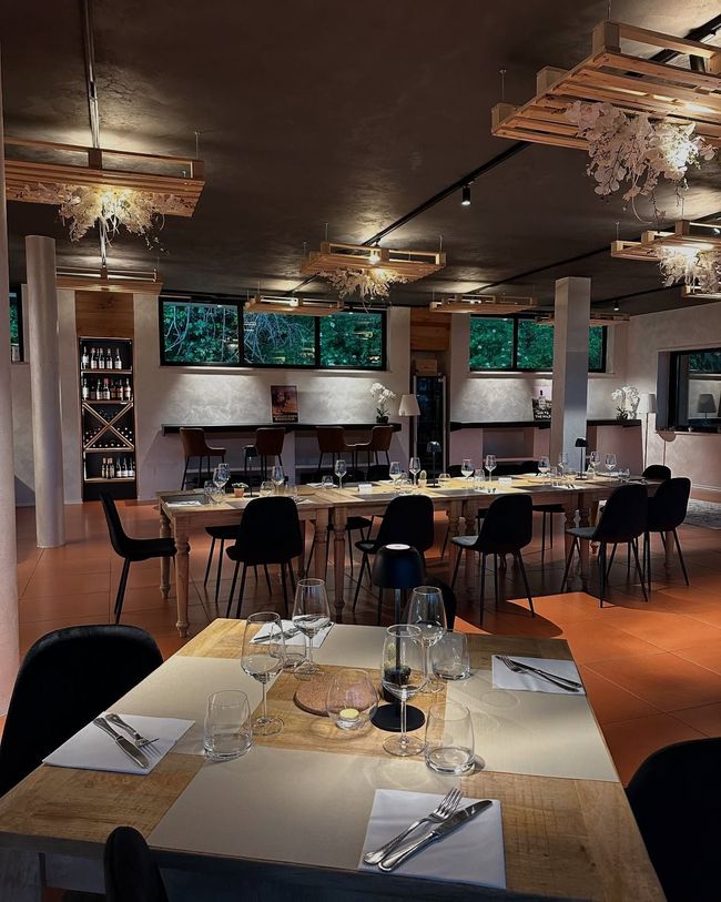 Interno del ristorante con lunghi tavoli in legno apparecchiati per cenare, sedie nere, luci soffuse e lampadari decorativi sul soffitto.