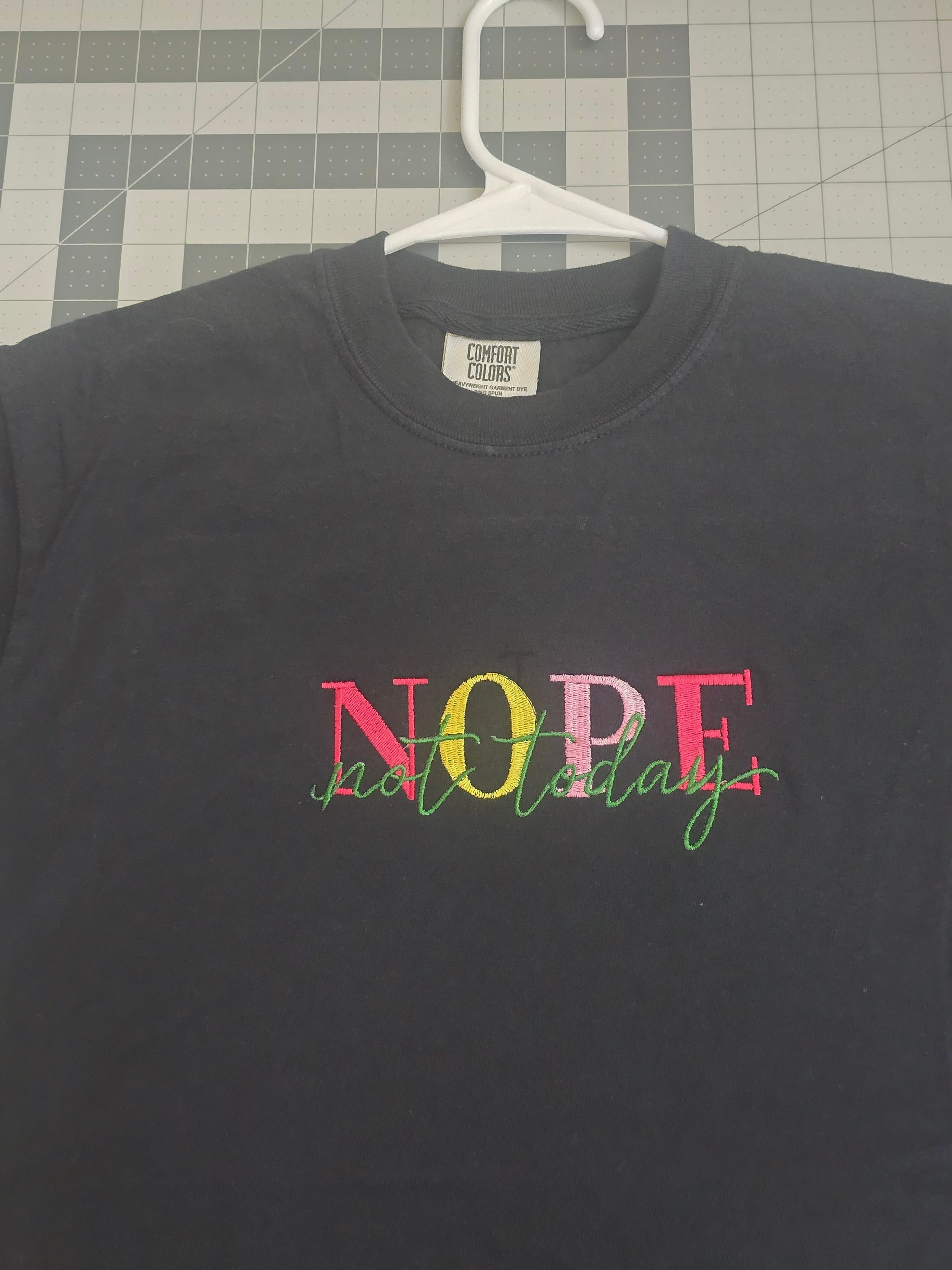 Woman in a black “NOPE” graphic T-shirt and jeans standing indoors beside a plant and sofa.