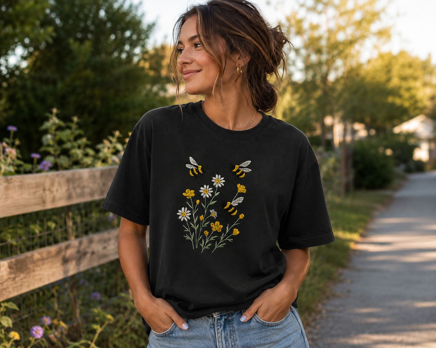 Woman in a black floral T-shirt standing by a wooden fence on a sunlit path, hands in pockets.