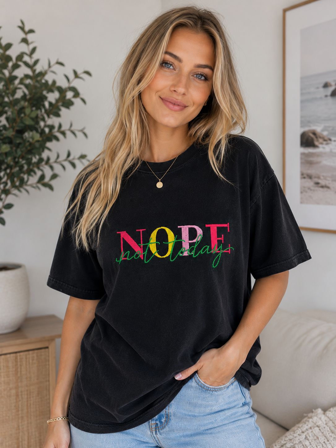 Woman in a black “NOPE” graphic T-shirt and jeans standing indoors beside a plant and sofa.