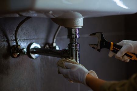 Gloved hands using a wrench to repair a sink pipe under a bathroom sink