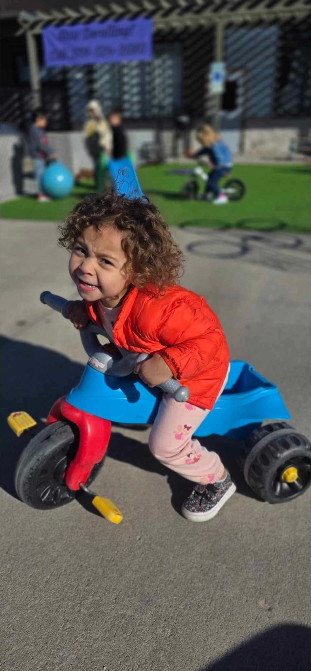 A child on a blue tricycle smiles, with red jacket and pink pants on asphalt, other kids in background.