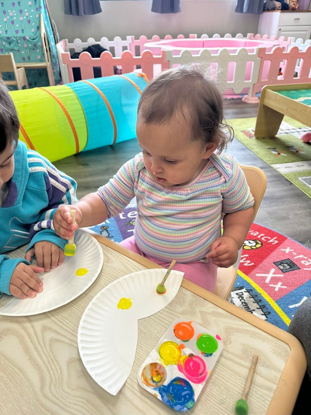 Child painting with yellow paint at a table, paper plates, paint palette.