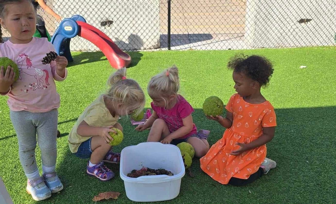 Children on green play surface examining objects, including a white bin.