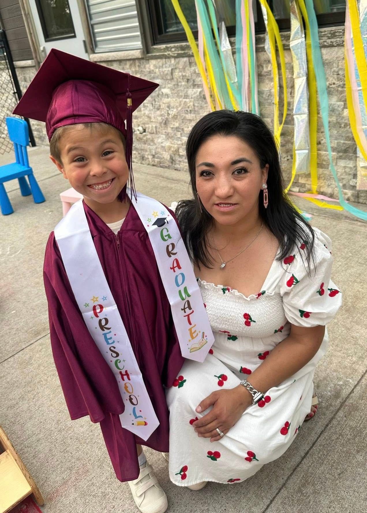 Child in graduation gown with woman, smiling outside. Colorful ribbons in background.