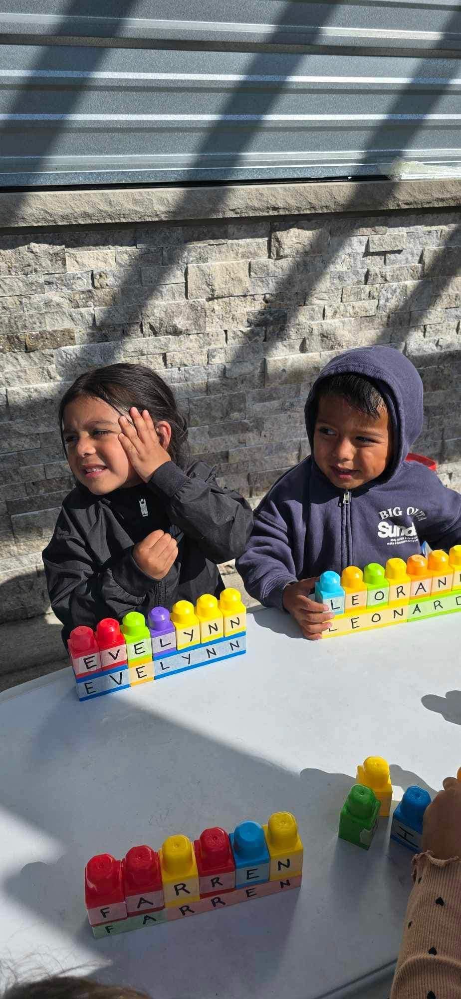 Two children playing with colorful blocks outdoors. One smiles, hand on face; the other in a hoodie smiles too.