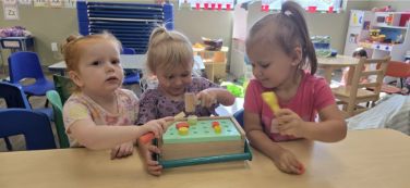 Three young children play at a table with a wooden toy. They use mallets and other tools, in a brightly lit room.