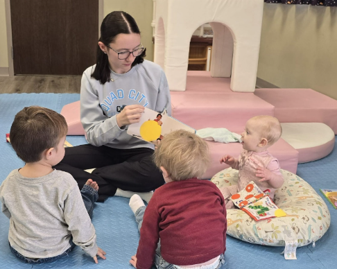 Woman reads a book to four children on a blue mat; a playhouse is in the background.