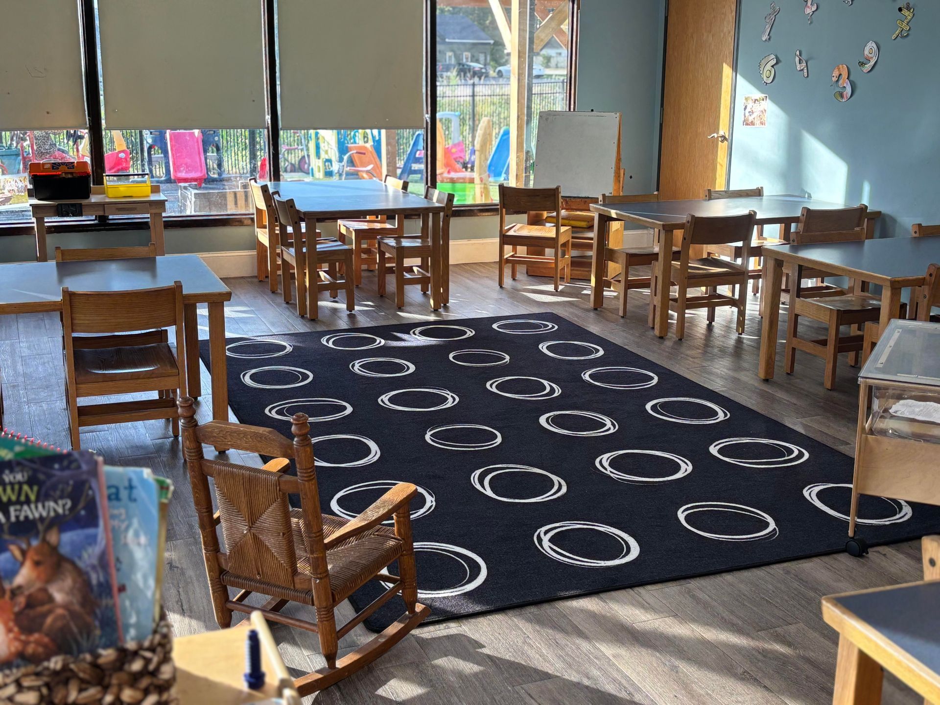 Classroom with tables, chairs, a rug, and a rocking chair. Sunlight streams in. Books on display.