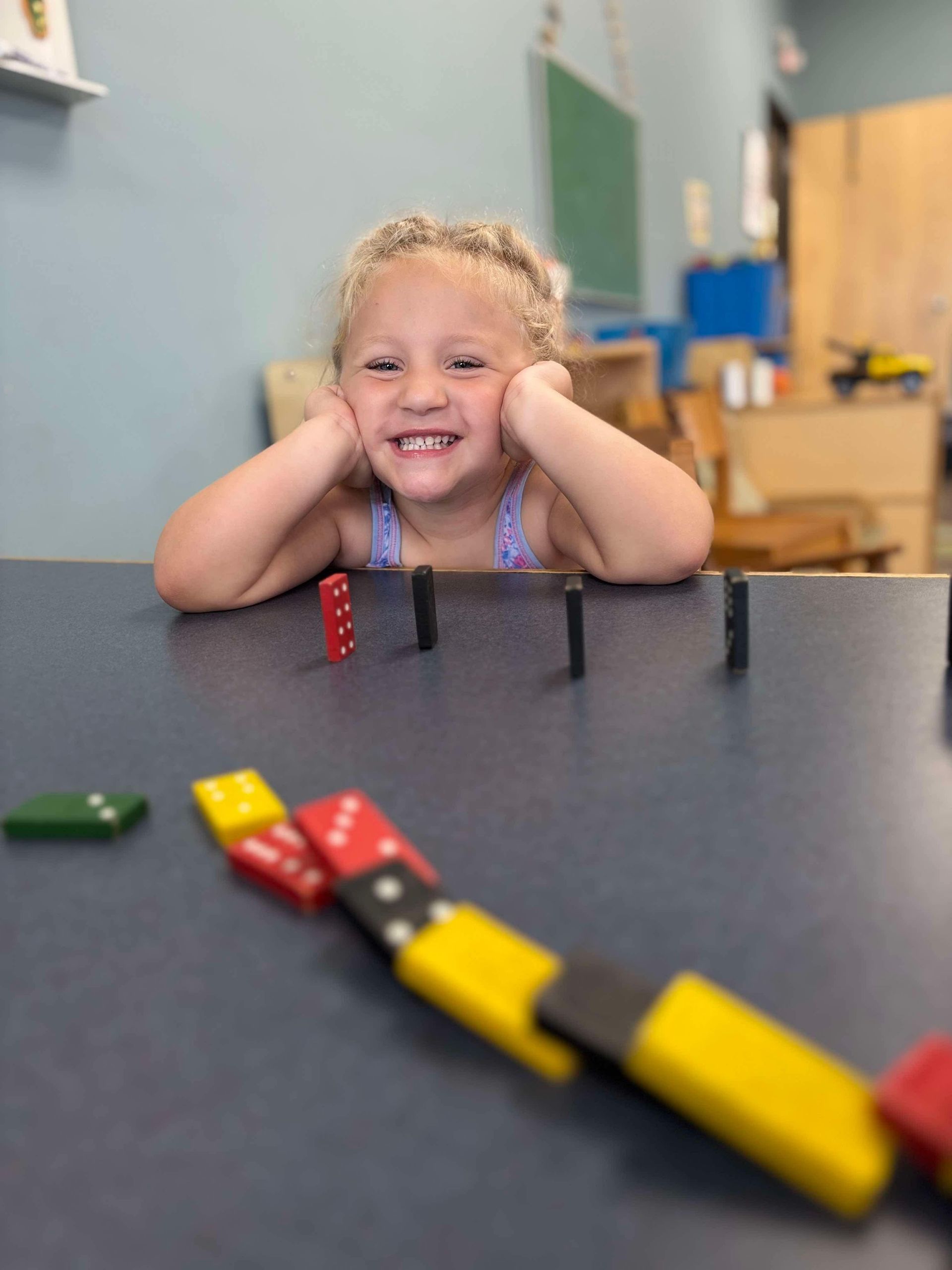 Smiling child watches dominoes on a table in a room.