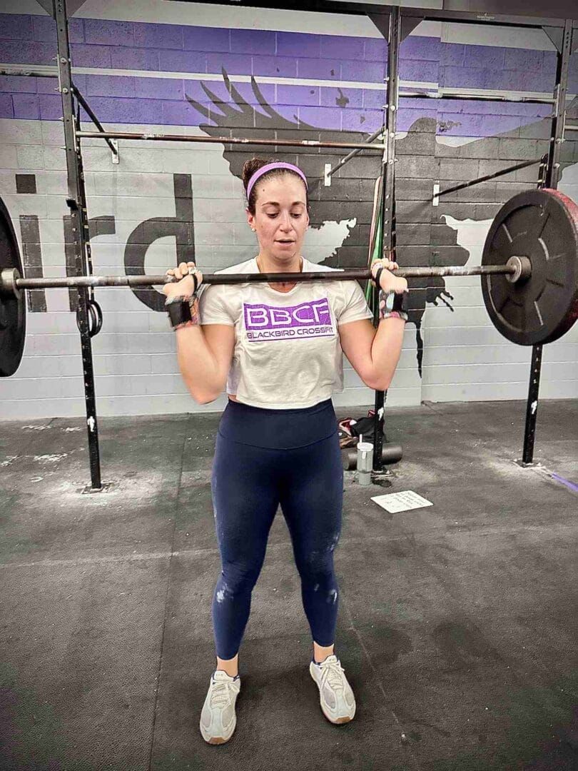 Person lifting a barbell in a gym, wearing a gray shirt, dark leggings, and white shoes.