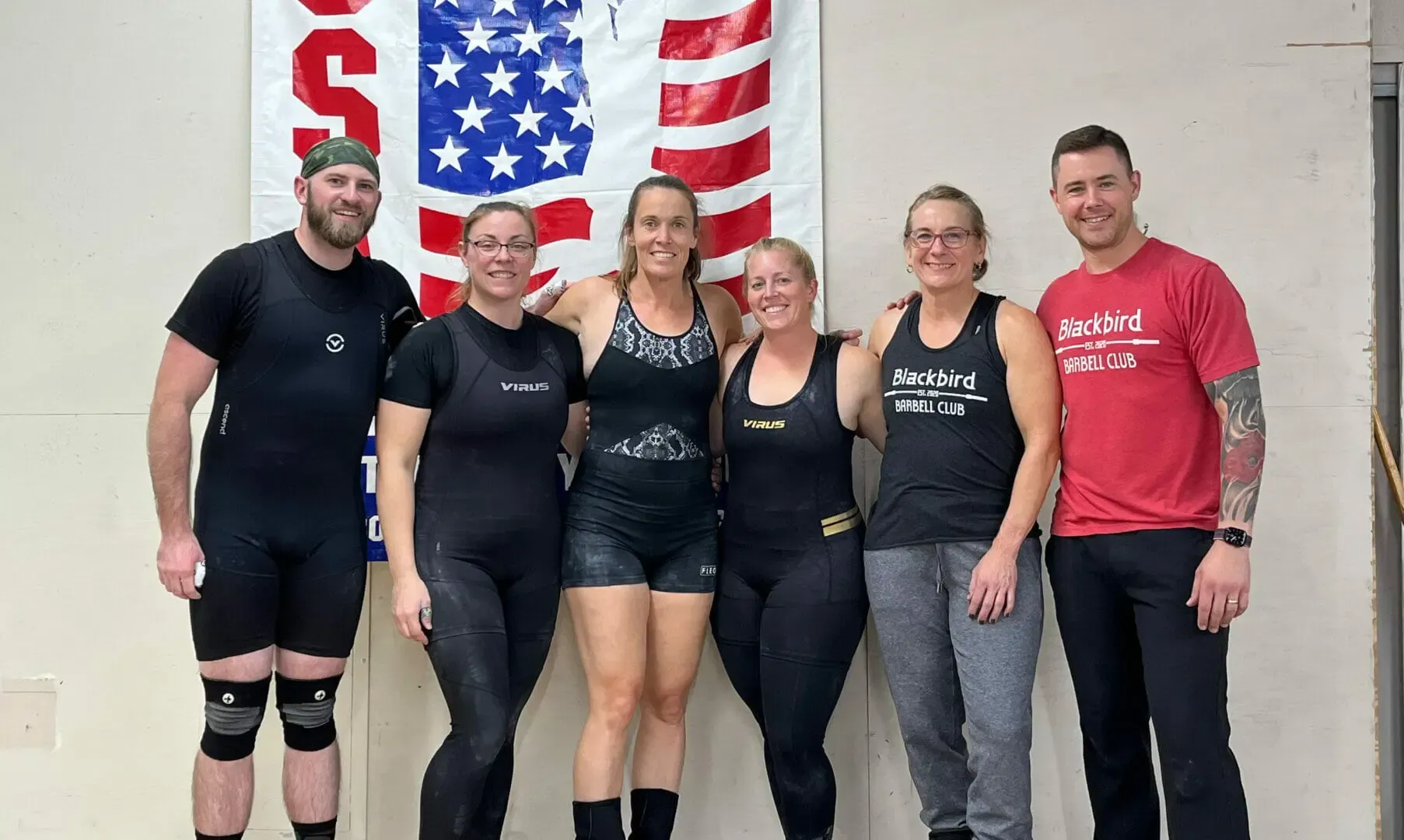 Five people posing indoors in front of a large U.S. flag, wearing workout clothes and smiling