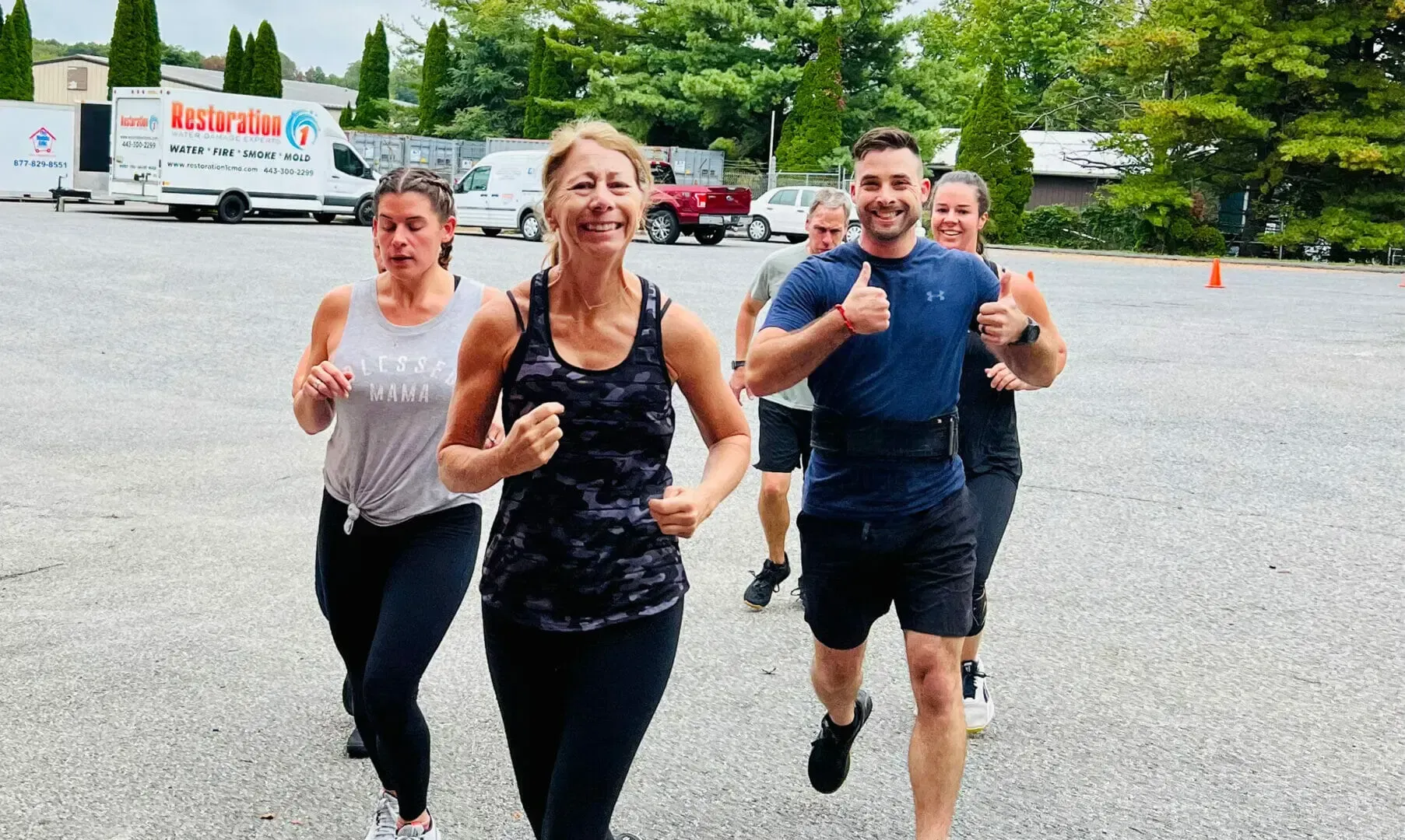 Four people jogging toward the camera on a paved lot, smiling and giving thumbs up.