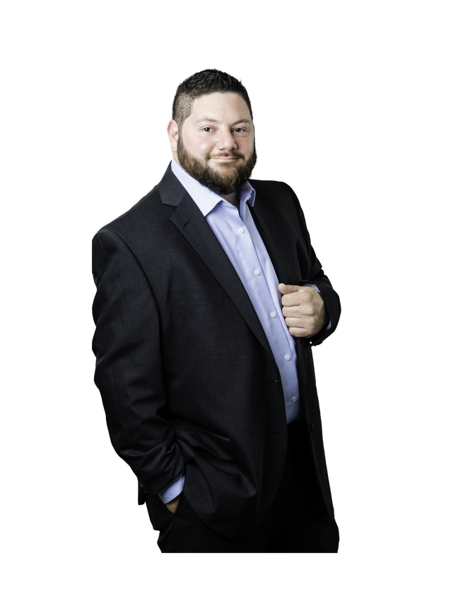 Man in a dark suit and light blue shirt standing against a white background, hand in pocket.