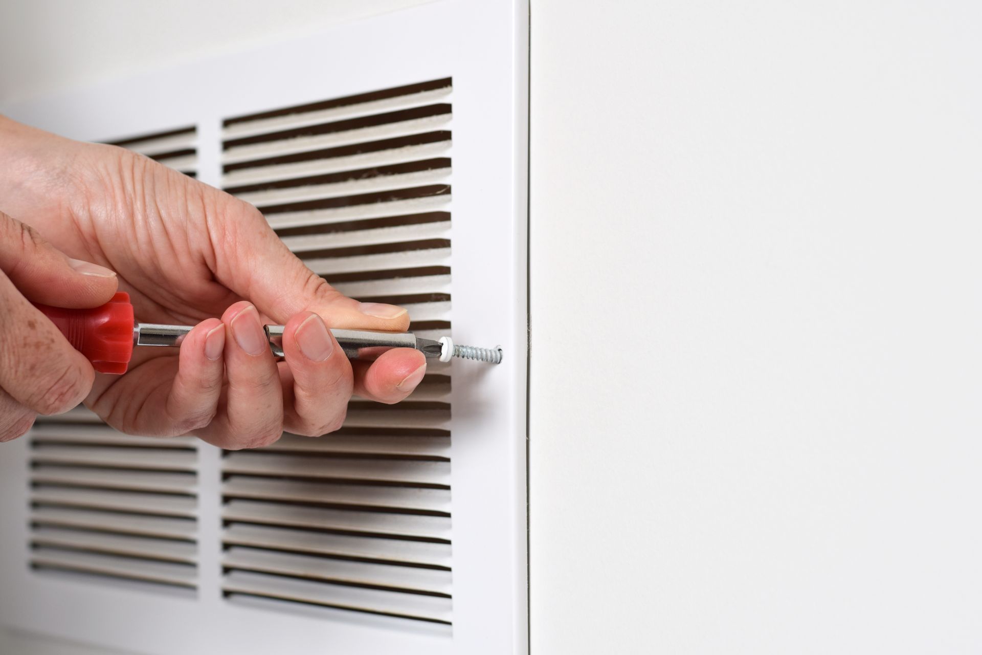 A person uses a red-handled screwdriver to secure a white wall vent cover.