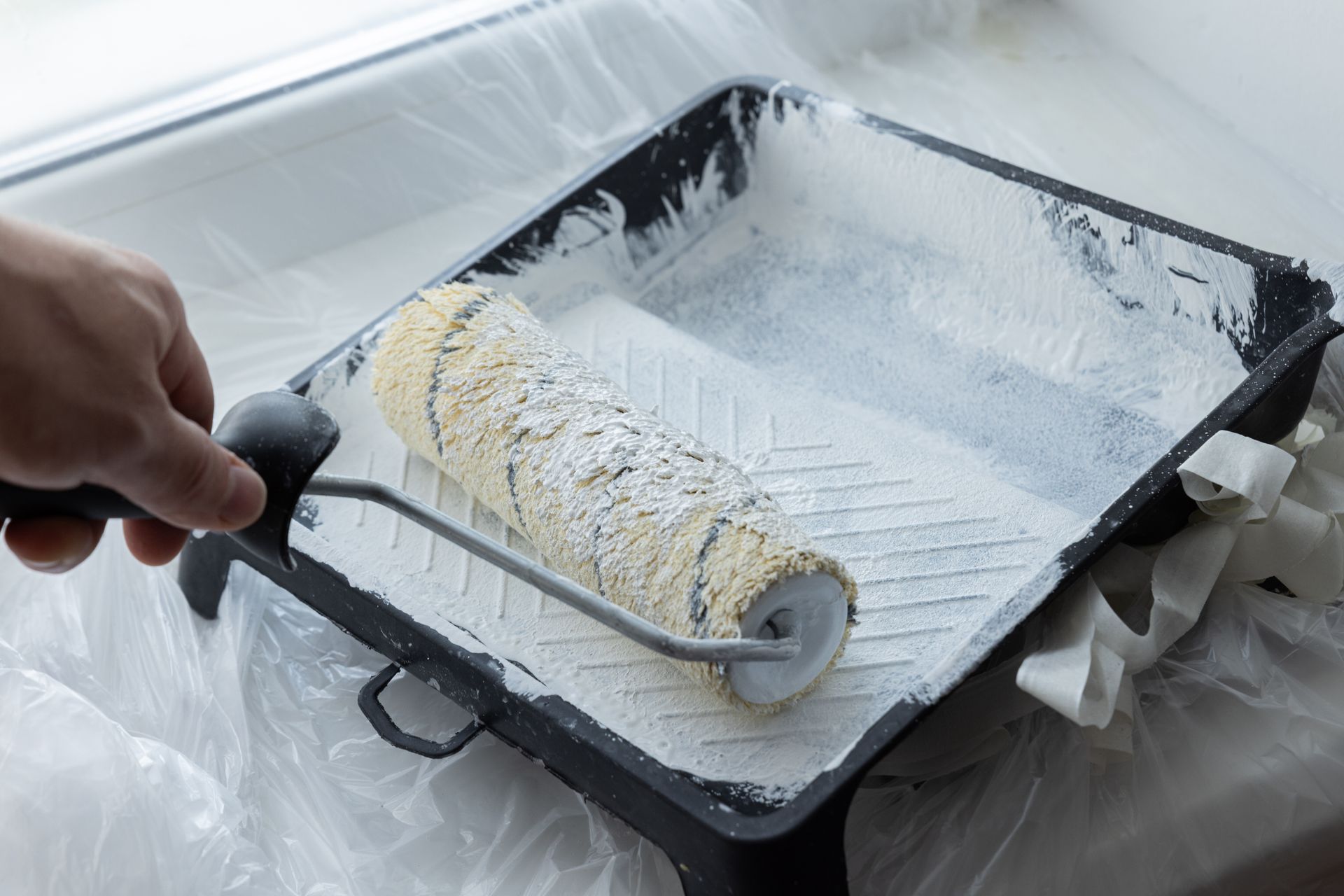 A hand holds a paint roller inside a black plastic tray coated with white paint.