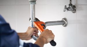 Plumber using an orange wrench to repair a silver pipe in a white tiled room.