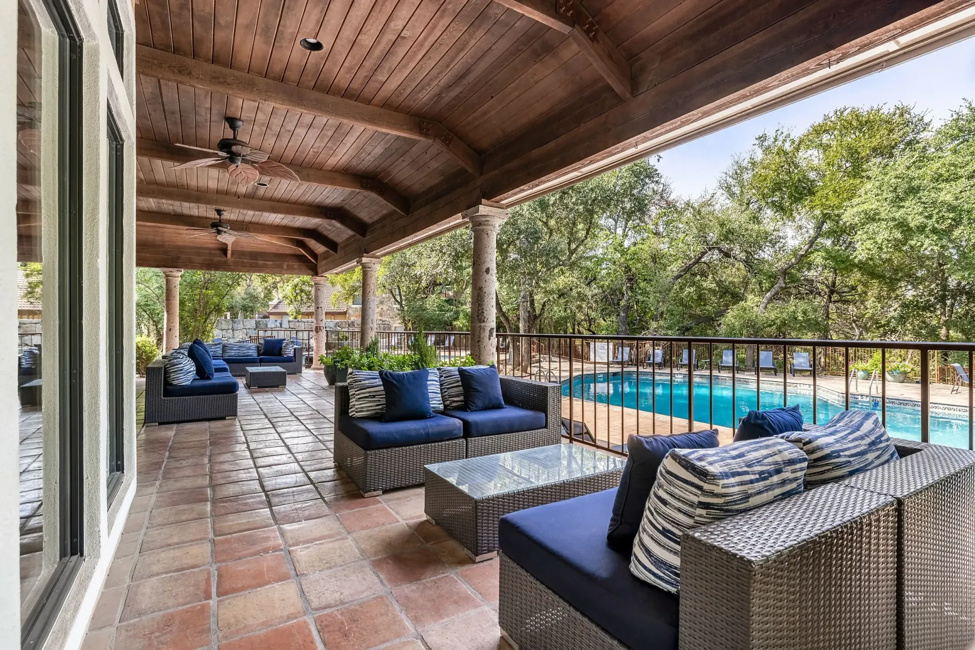 Covered outdoor patio with comfortable seating overlooking a swimming pool and lush trees.