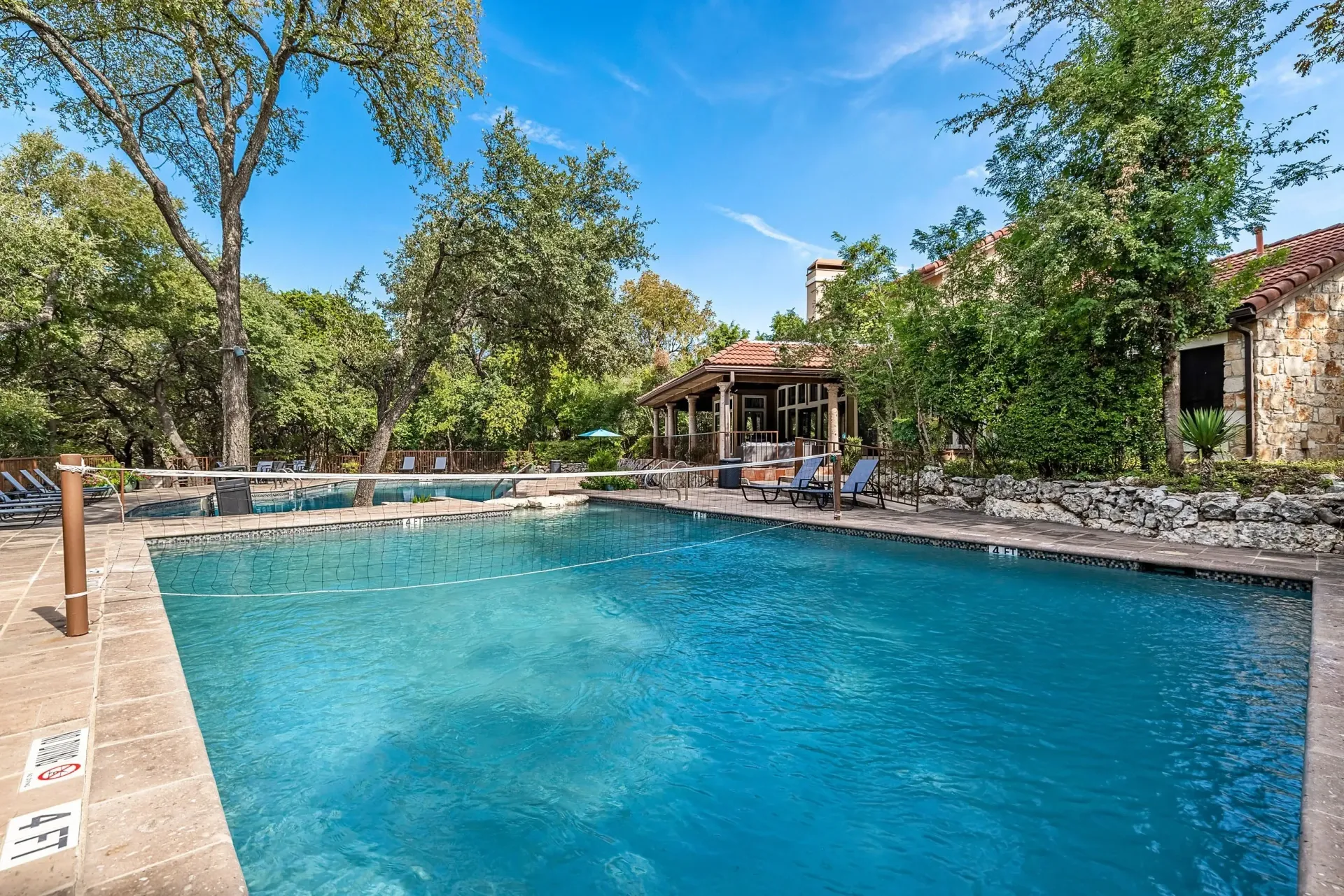 Outdoor swimming pool with a volleyball net and lounge chairs, surrounded by trees and a building.