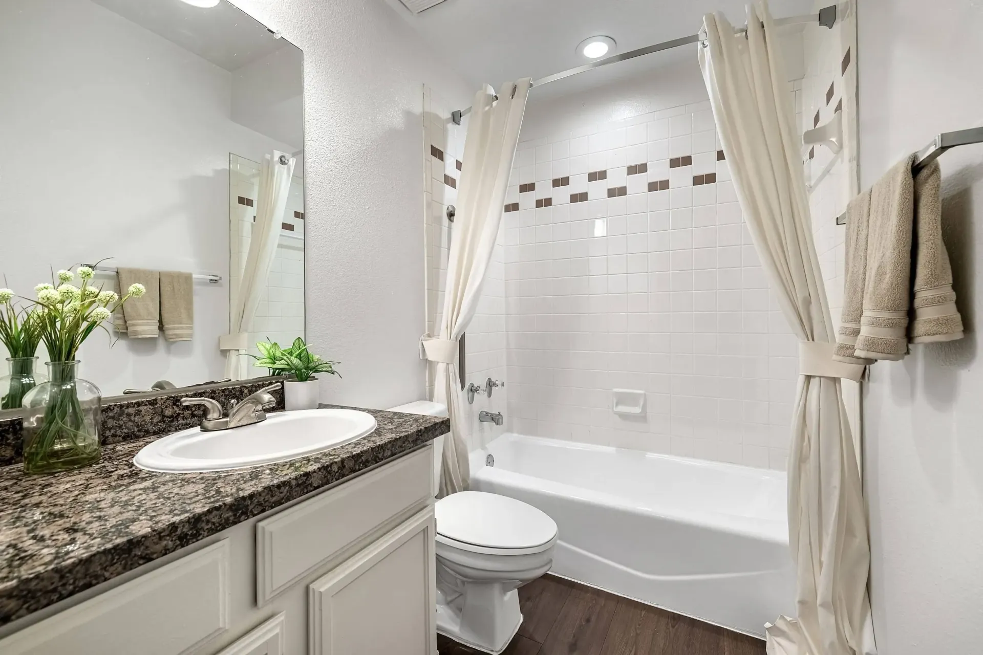 A bright and clean bathroom featuring a granite countertop vanity with a white sink, a toilet, and a white tiled shower/tub combination with a cream-colored shower curtain.