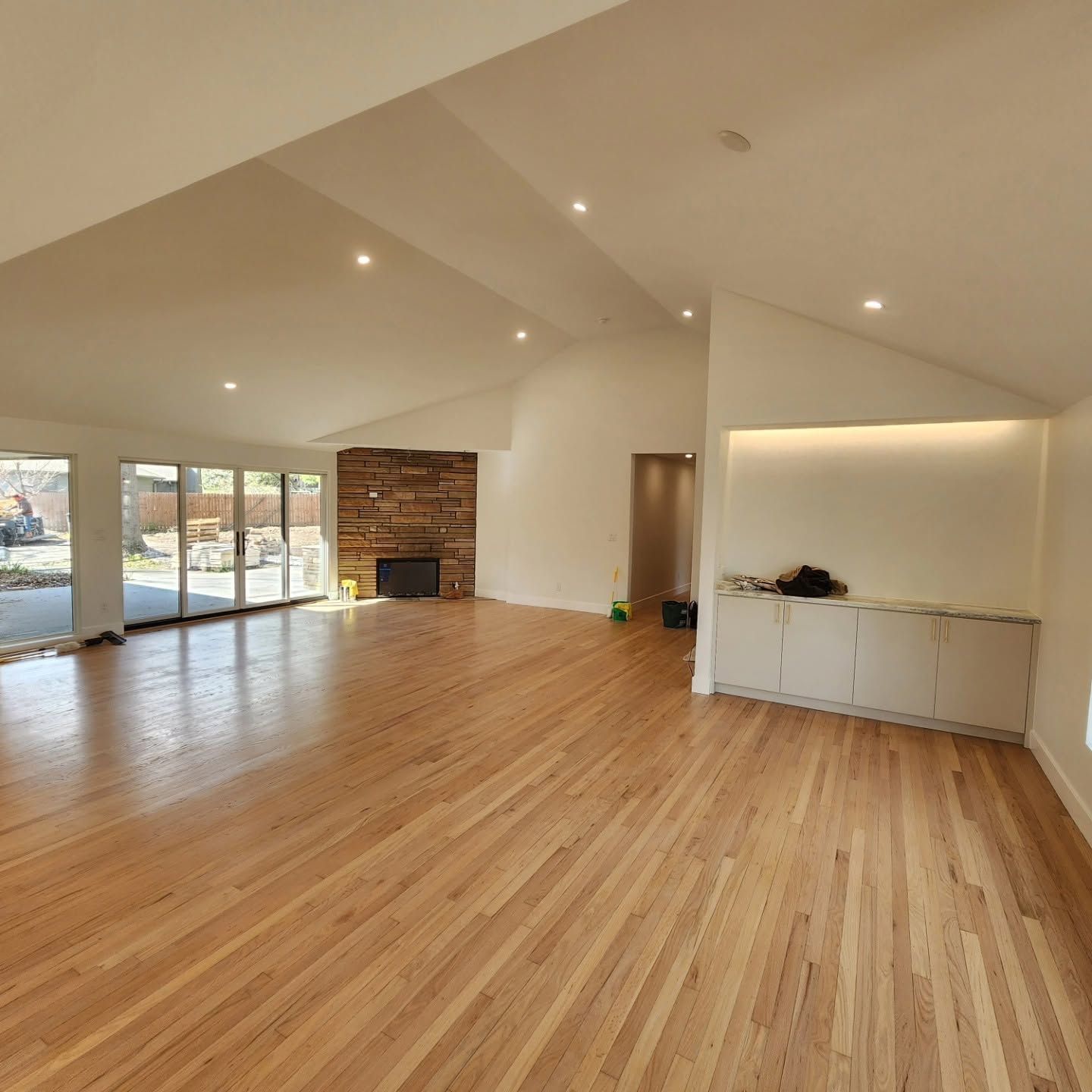 Empty modern living room with vaulted ceilings, hardwood floors, large windows, and built-in cabinetry.
