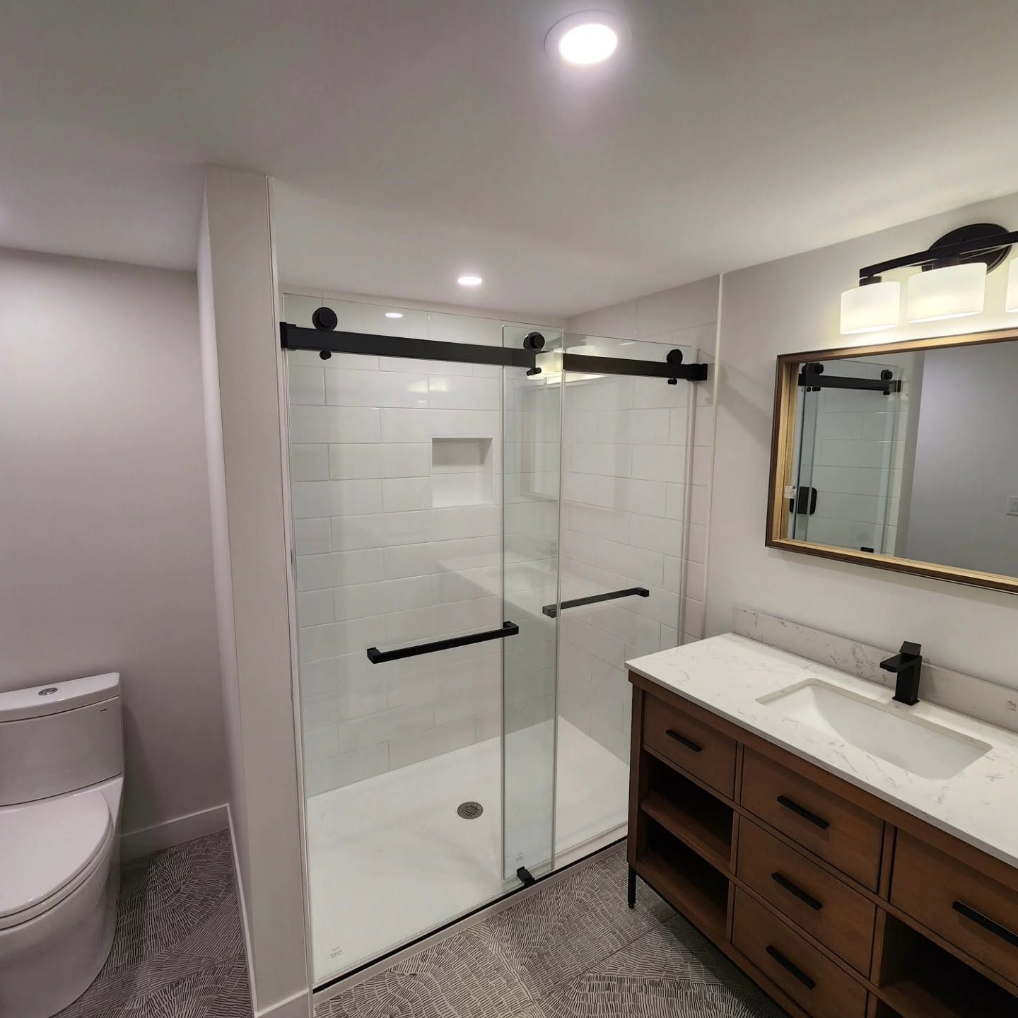 Modern bathroom with glass shower, white tiles, toilet, and wooden vanity with mirror.