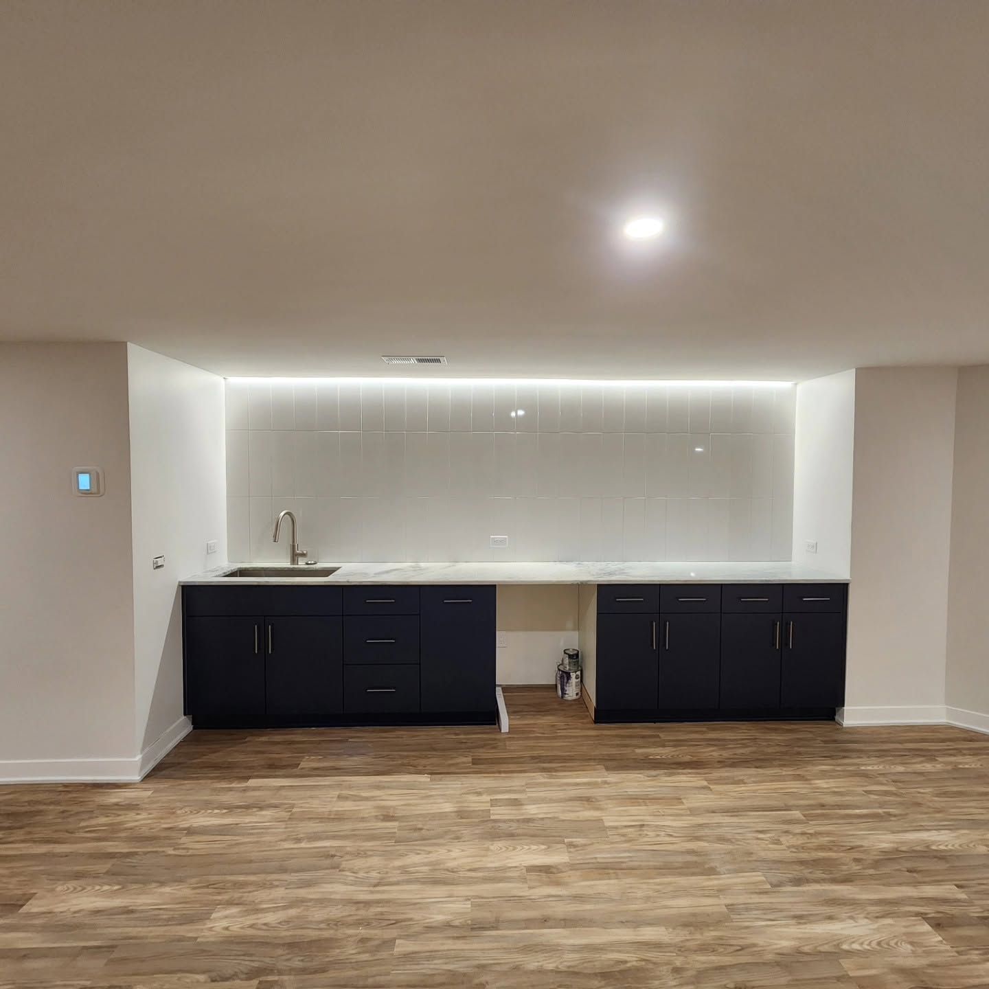 Empty modern kitchen with dark cabinets, white backsplash, and wood flooring in a finished basement