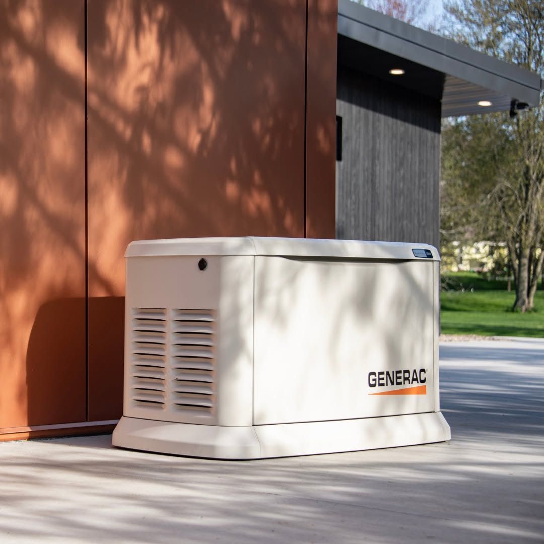 White Generac generator beside a brown building on a concrete driveway, with trees in the background