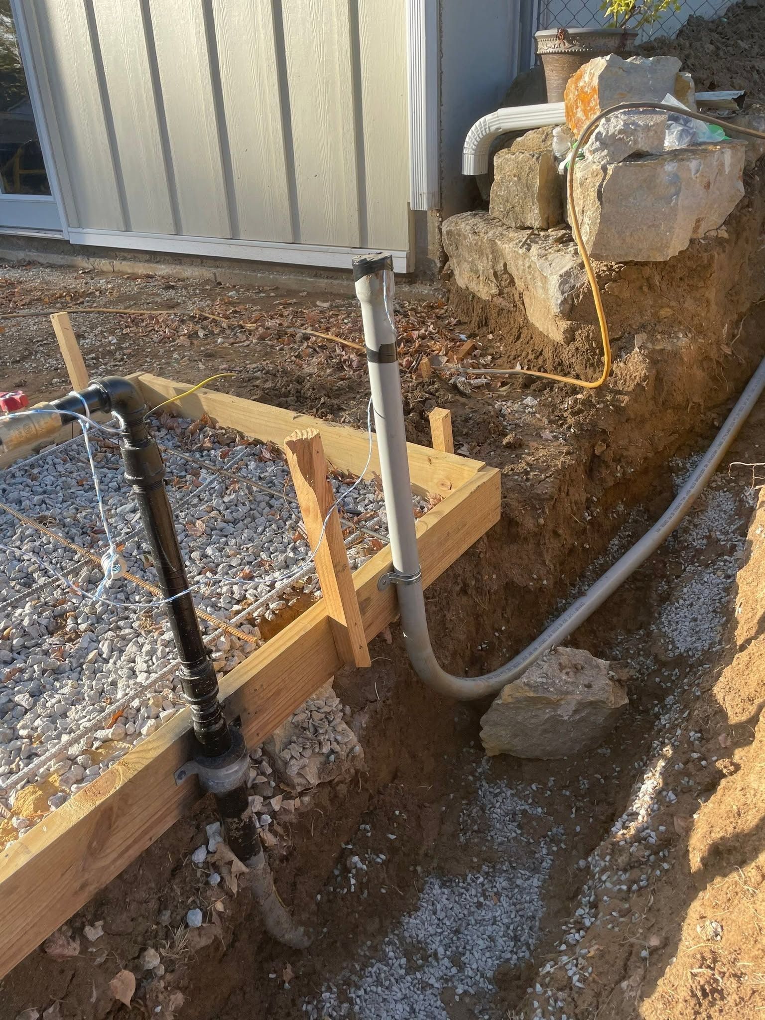 Trenched utility pipes beside a metal building, with gravel, dirt, and a concrete footing.