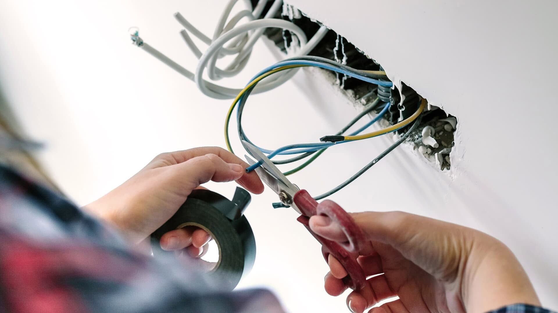 Hands using red-handled pliers to wire a ceiling electrical box with exposed cables.