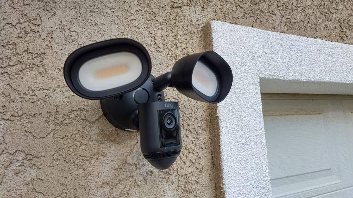 Wall-mounted black security light camera beside a white exterior window on a stucco wall