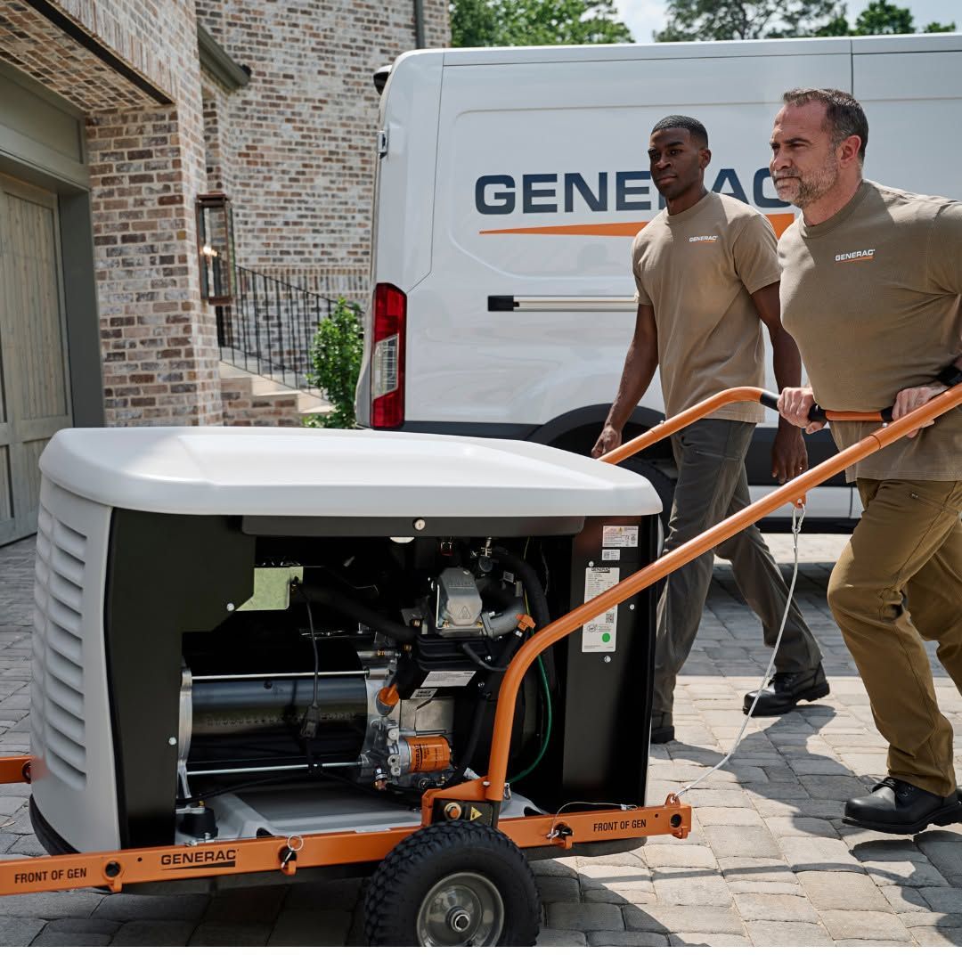 Two workers moving a portable generator on a wheeled cart beside a white van in a driveway.