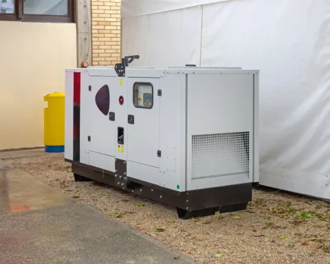Large gray industrial generator unit beside a white building on gravel