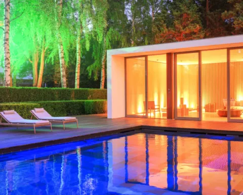 Lit pool beside a modern villa at night, with warm interior lights and two lounge chairs by the hedge
